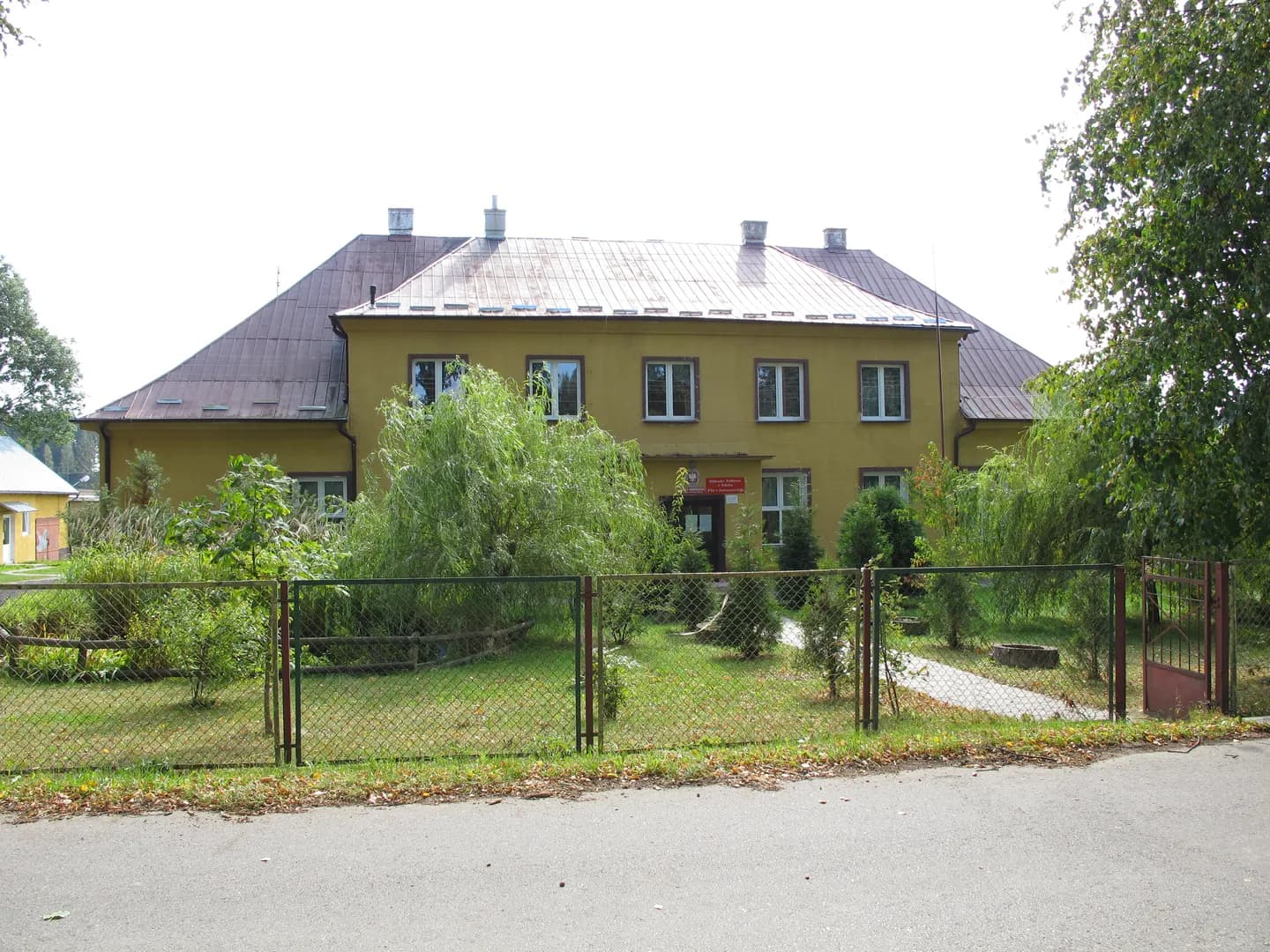 Budynek Szkoły Podstawowej w Janowszczyźnie z ogrodem i ogrodzeniem na pierwszym planie.