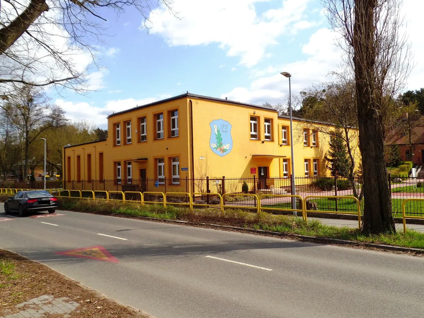 Żółty budynek Szkoły Podstawowej nr 17 w Toruniu z herbem na elewacji widoczny od strony ulicy