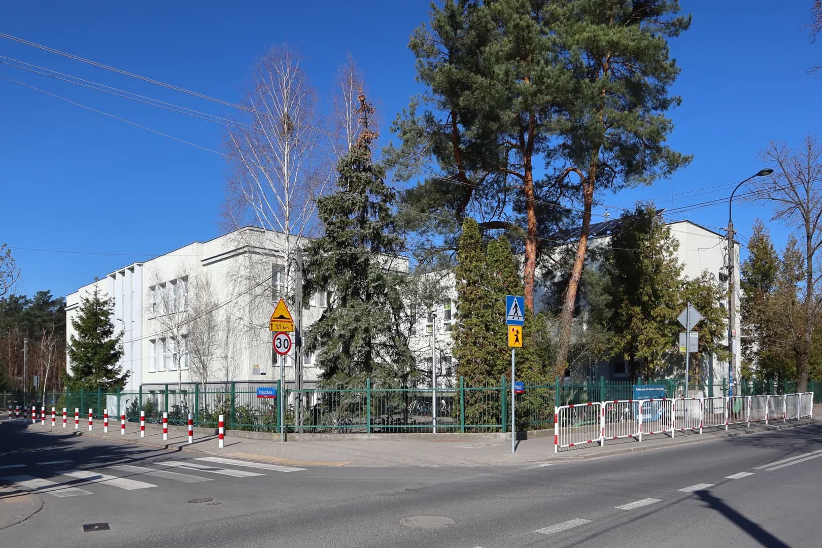 Widok na budynek Szkoły Podstawowej nr 76 w Warszawie zza ogrodzenia i drzew