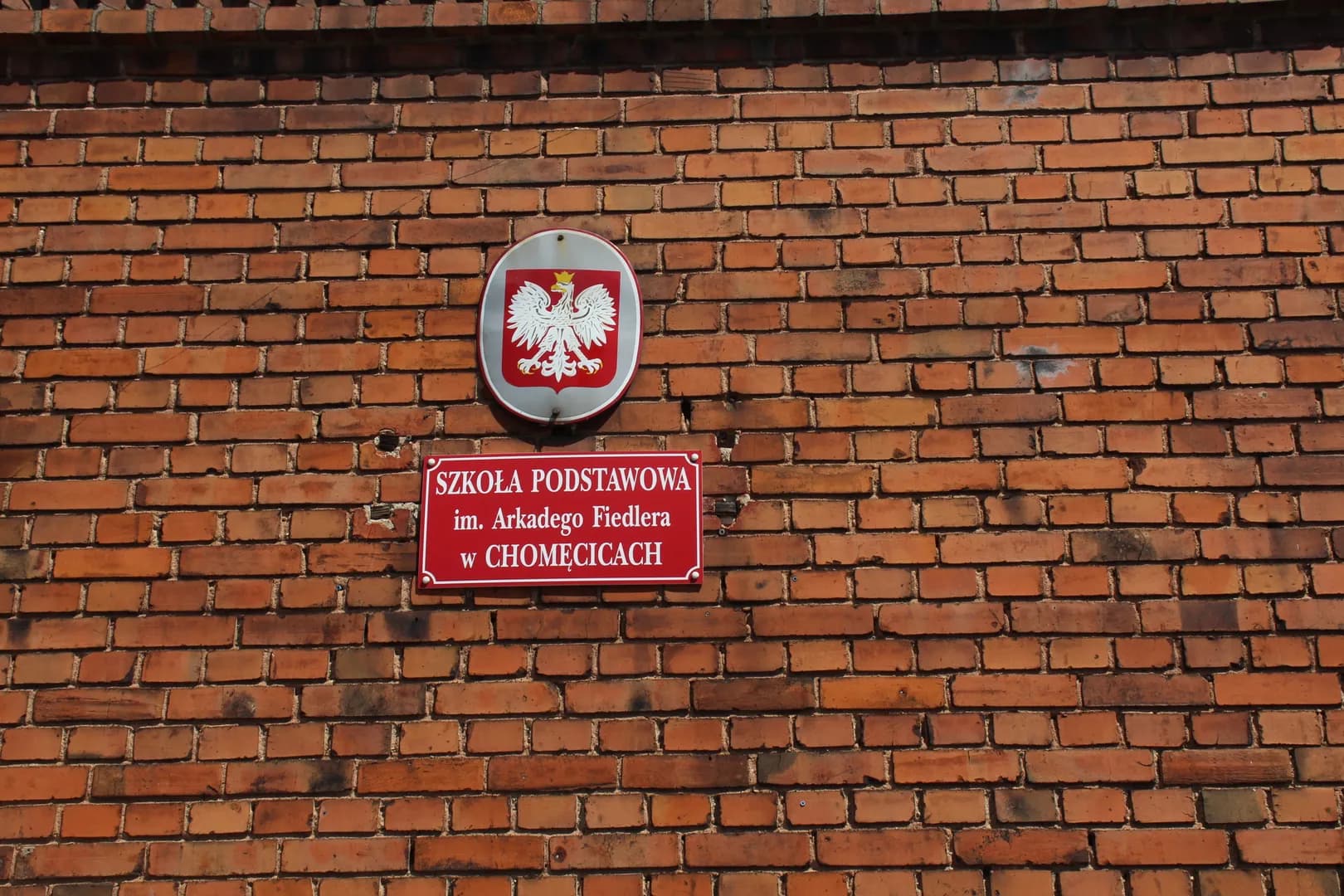 Tablica informacyjna Szkoły Podstawowej im. Arkadego Fiedlera w Chomęcicach na ceglanym murze.