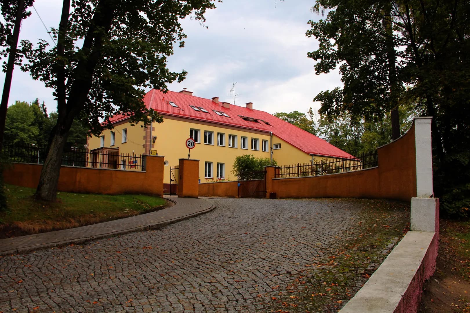 Budynek szkoły z żółtą elewacją i czerwonym dachem za brukowanym podjazdem